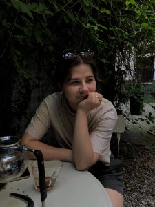 dziewczyna w jasnej koszulce, siedząca w ogrodzie przy kawie, zapatrzona w dal, na stole znajduje się przyrząd do przygotowania kawy oraz filiżanka