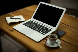 biała filiżanka kawy na drewnianym stole, obok komputer- MacBook firmy Apple