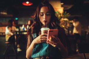 Młoda dziewczyna w kawiarni, pijąca kawę mrożoną w plastikowym, jednorazowym kubku, patrząca w telefon z różową obudową